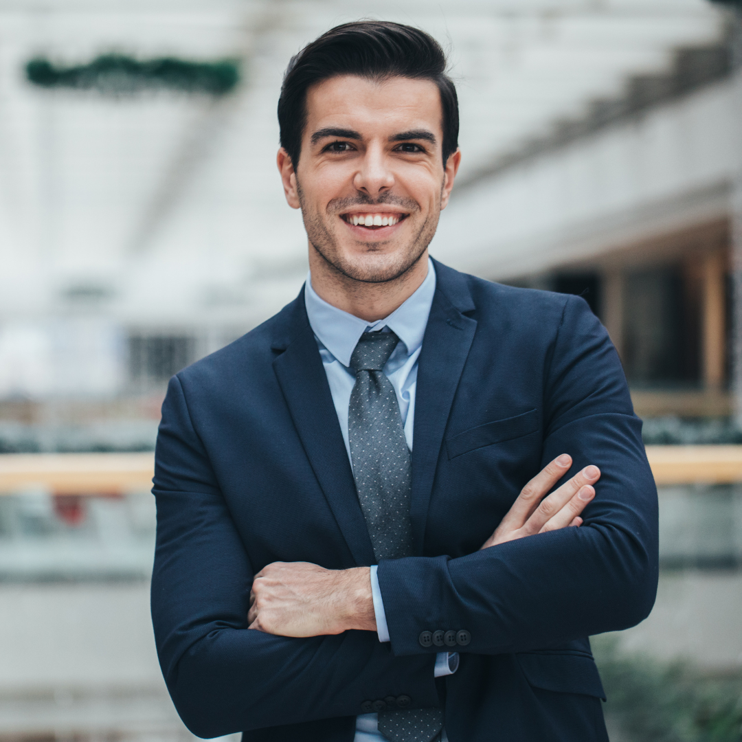 Hombre joven con traje azul marino y corbata gris, sonriendo con los brazos cruzados en un entorno empresarial moderno.